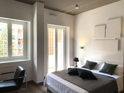 a bedroom with a bed with a chair and two windows at Nox Romana Apartments - Vatican - Modern Apt with Cozy Balcony in Rome